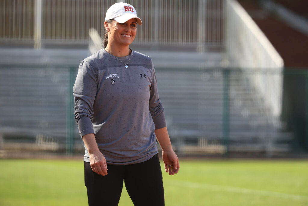 Photo: A white woman in casual attire struts, with a smile, onto a softball field. She wears a white cap with BU's logo on the front.