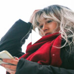 Photo: A young woman holds her head in her hand as she looks down at her phone. She has dyed blonde hair and wears a read scarf around her neck. Play button overlay.