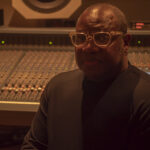 Photo: A photo with a older Black man sitting in a recording studio with the dials and controls behind him.